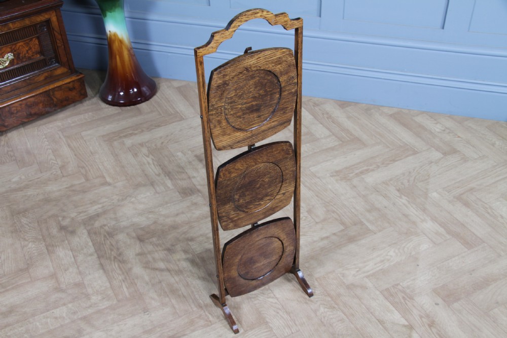 An Edwardian solid oak three-tier folding cake stand featuring square tiered shelves with recessed circular plate holders and an arched top handle. View IV