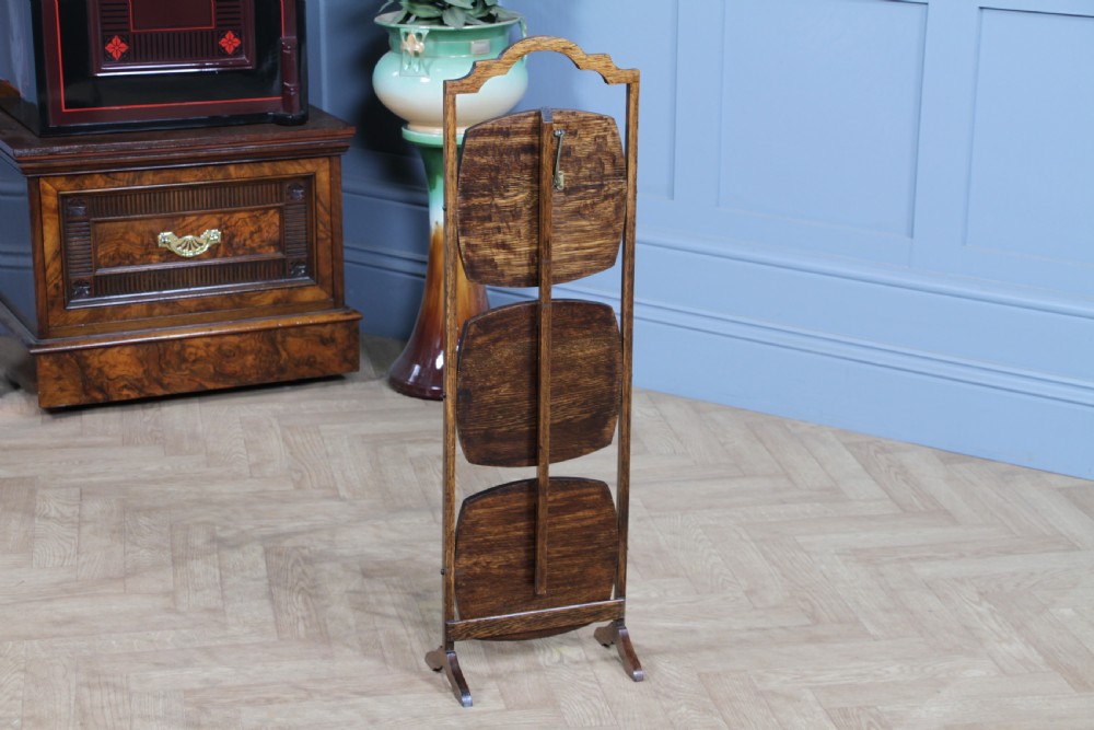 An Edwardian solid oak three-tier folding cake stand featuring square tiered shelves with recessed circular plate holders and an arched top handle. View X