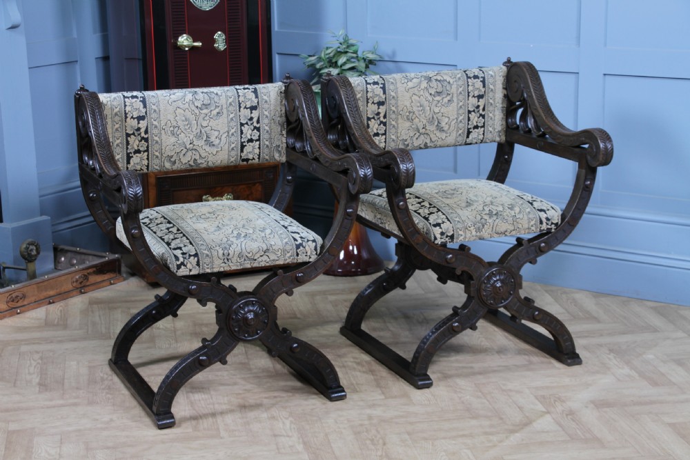 A pair of carved dark oak x-frame curule armchairs featuring patterned floral upholstery and central carved rosette bosses on the legs.