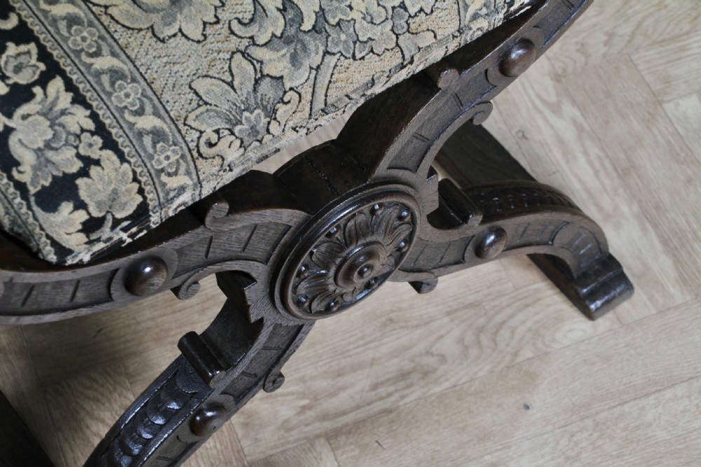A pair of carved dark oak x-frame curule armchairs featuring patterned floral upholstery and central carved rosette bosses on the legs. View IX