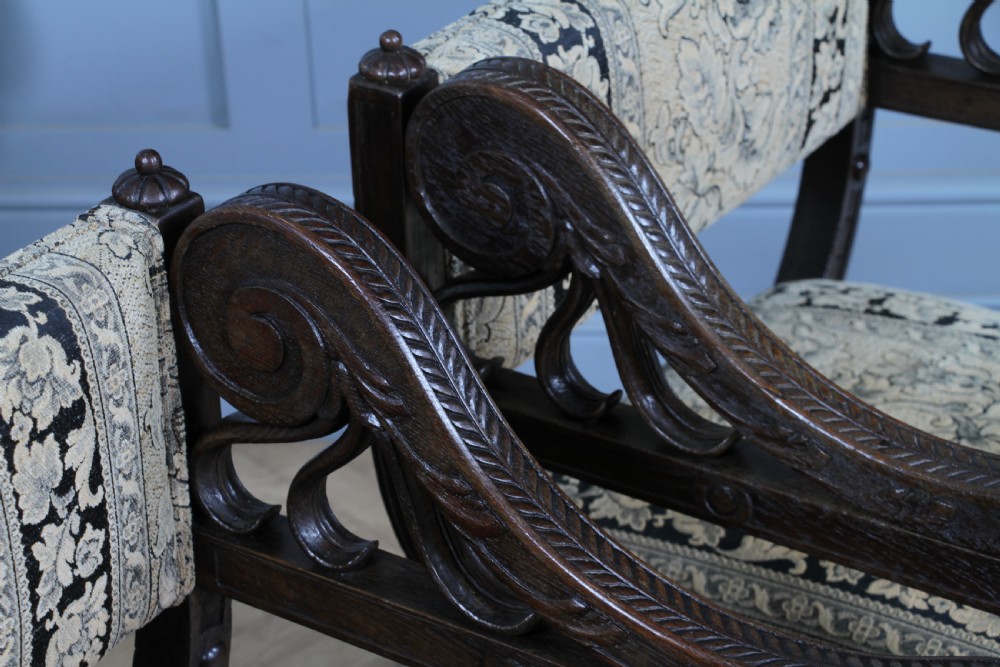 A pair of carved dark oak x-frame curule armchairs featuring patterned floral upholstery and central carved rosette bosses on the legs. View X