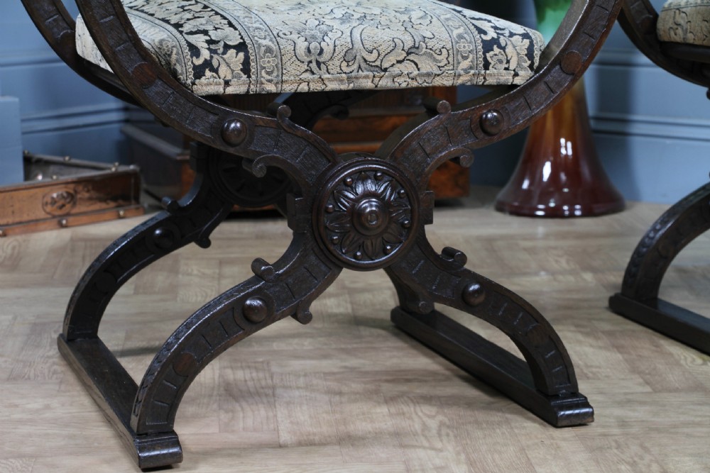 A pair of carved dark oak x-frame curule armchairs featuring patterned floral upholstery and central carved rosette bosses on the legs. View XII