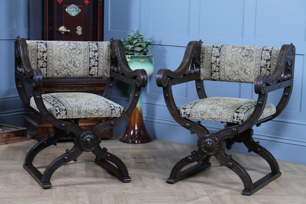 A pair of carved dark oak x-frame curule armchairs featuring patterned floral upholstery and central carved rosette bosses on the legs. View XIV