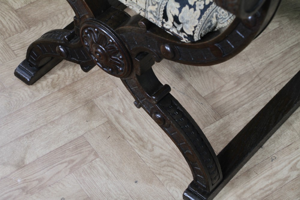 A pair of carved dark oak x-frame curule armchairs featuring patterned floral upholstery and central carved rosette bosses on the legs. View XV
