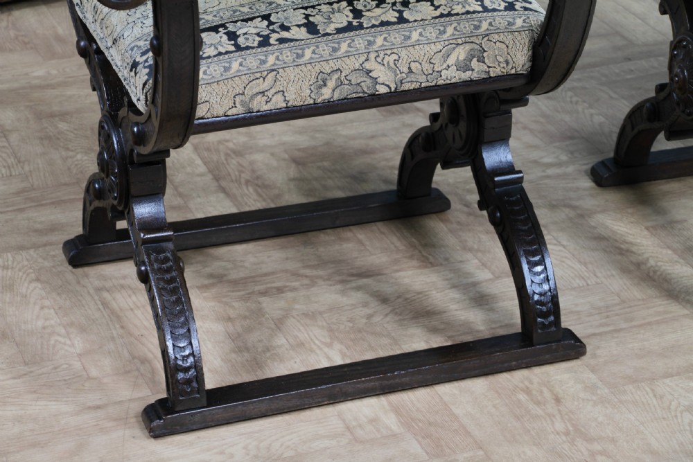 A pair of carved dark oak x-frame curule armchairs featuring patterned floral upholstery and central carved rosette bosses on the legs. View XVI