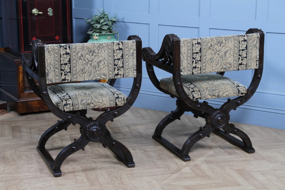 A pair of carved dark oak x-frame curule armchairs featuring patterned floral upholstery and central carved rosette bosses on the legs. View XIX