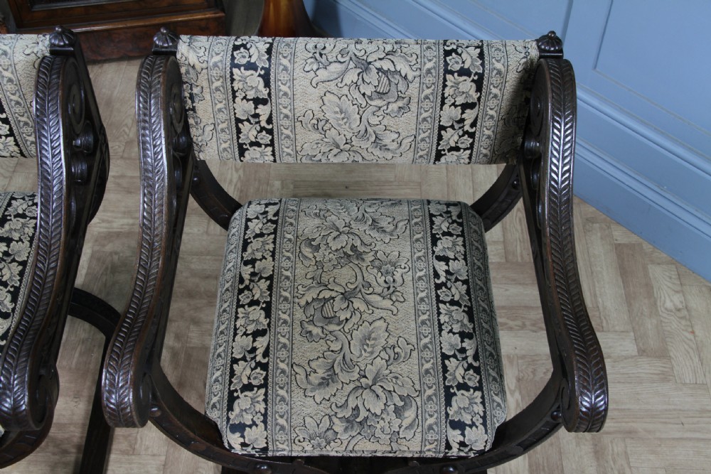 A pair of carved dark oak x-frame curule armchairs featuring patterned floral upholstery and central carved rosette bosses on the legs. View XXI