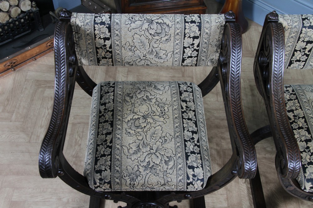 A pair of carved dark oak x-frame curule armchairs featuring patterned floral upholstery and central carved rosette bosses on the legs. View XXIII