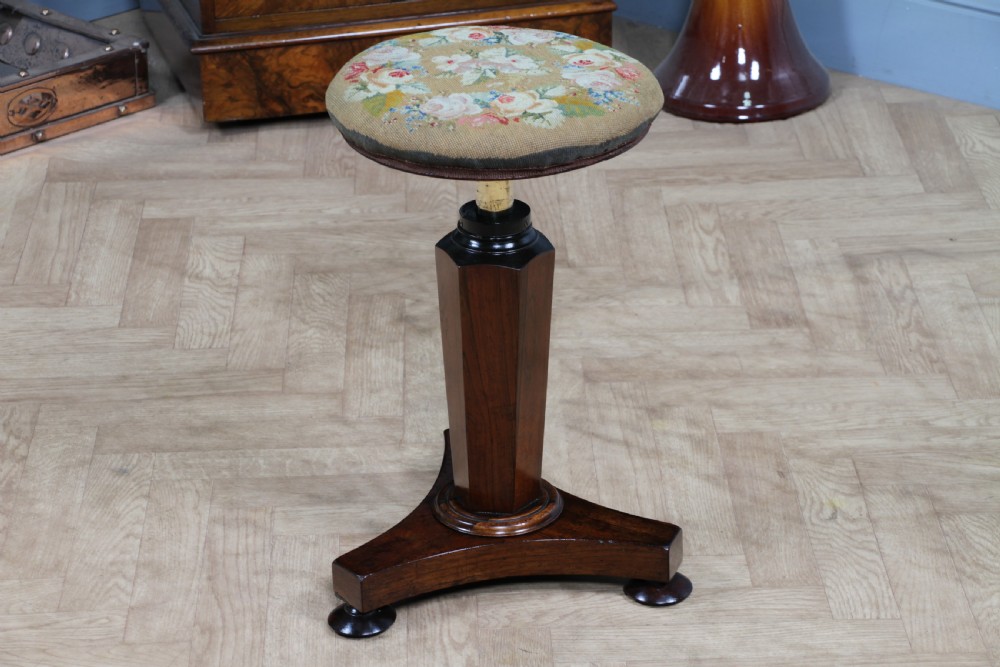 An early Victorian rosewood revolving piano stool featuring a circular floral needlepoint seat, a faceted central column, and a solid triform base with turned feet.