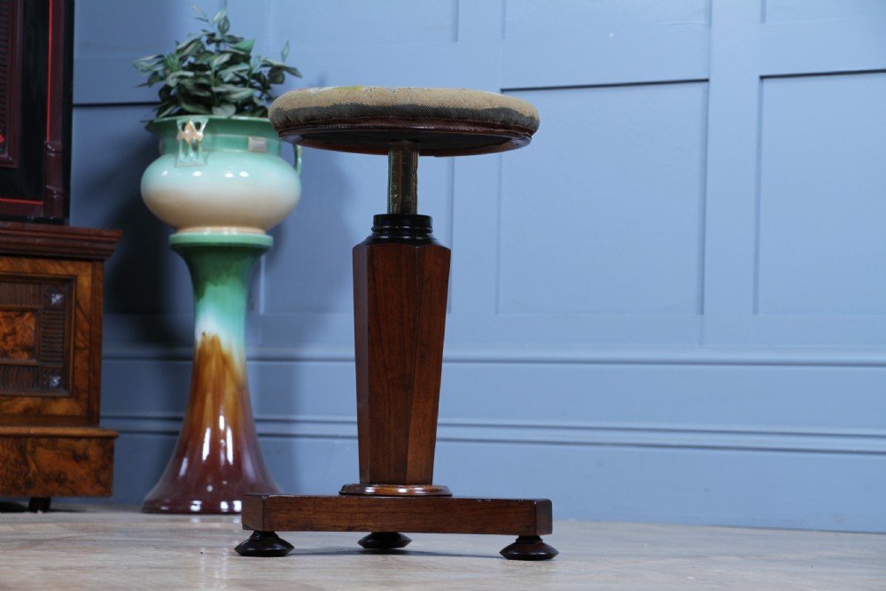 An early Victorian rosewood revolving piano stool featuring a circular floral needlepoint seat, a faceted central column, and a solid triform base with turned feet. View IV