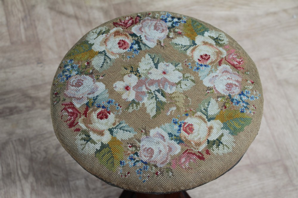 An early Victorian rosewood revolving piano stool featuring a circular floral needlepoint seat, a faceted central column, and a solid triform base with turned feet. View V
