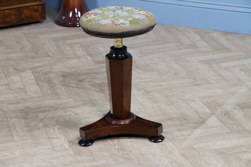 An early Victorian rosewood revolving piano stool featuring a circular floral needlepoint seat, a faceted central column, and a solid triform base with turned feet. View VII