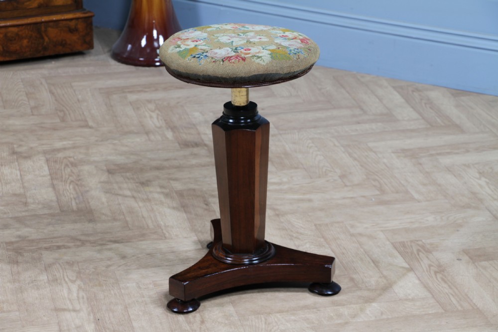 An early Victorian rosewood revolving piano stool featuring a circular floral needlepoint seat, a faceted central column, and a solid triform base with turned feet. View XIII