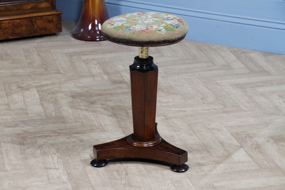 An early Victorian rosewood revolving piano stool featuring a circular floral needlepoint seat, a faceted central column, and a solid triform base with turned feet. View XIV