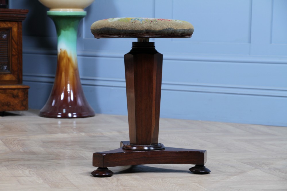 An early Victorian rosewood revolving piano stool featuring a circular floral needlepoint seat, a faceted central column, and a solid triform base with turned feet. View XV