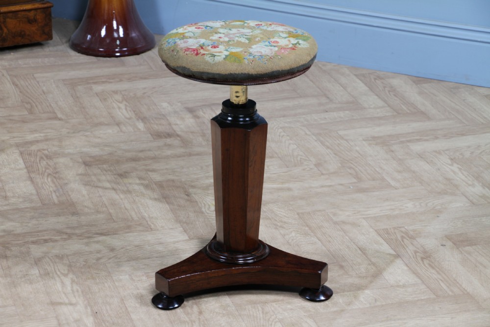 An early Victorian rosewood revolving piano stool featuring a circular floral needlepoint seat, a faceted central column, and a solid triform base with turned feet. View XVII