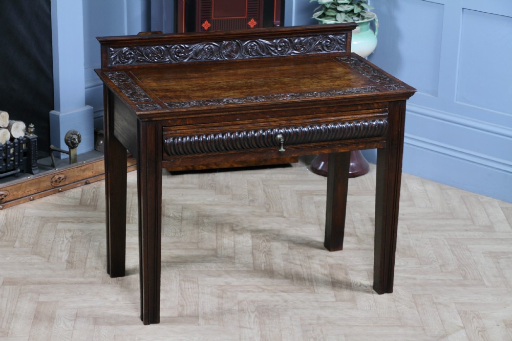 A dark carved oak side desk featuring a carved low gallery back, border-carved top surface, a single fluted drawer, and reeded legs.