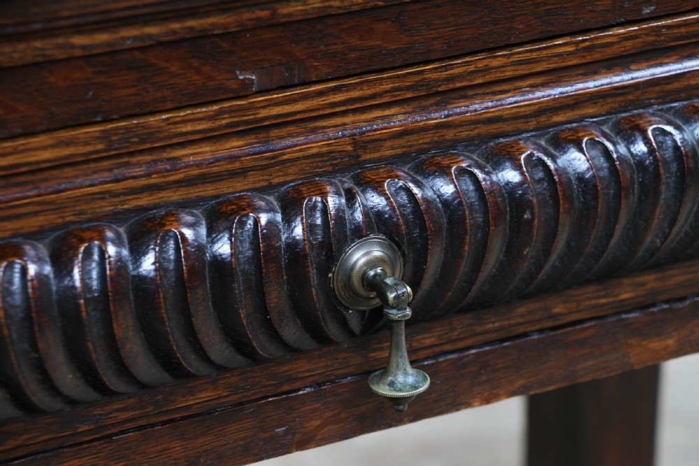 A dark carved oak side desk featuring a carved low gallery back, border-carved top surface, a single fluted drawer, and reeded legs. View VII