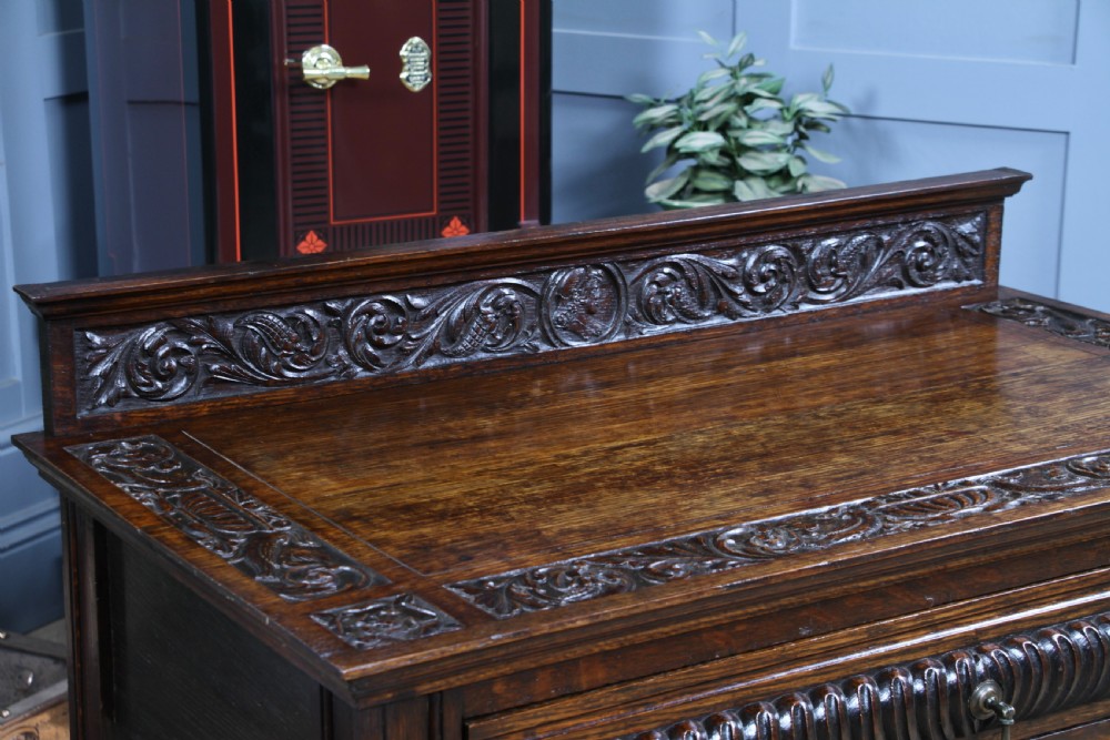A dark carved oak side desk featuring a carved low gallery back, border-carved top surface, a single fluted drawer, and reeded legs. View IX
