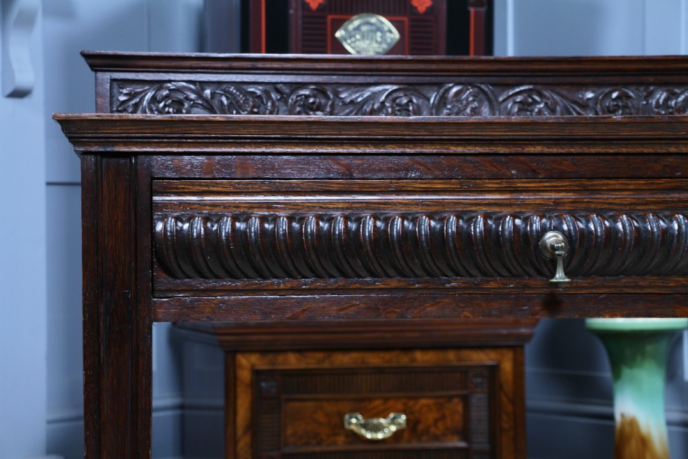 A dark carved oak side desk featuring a carved low gallery back, border-carved top surface, a single fluted drawer, and reeded legs. View X