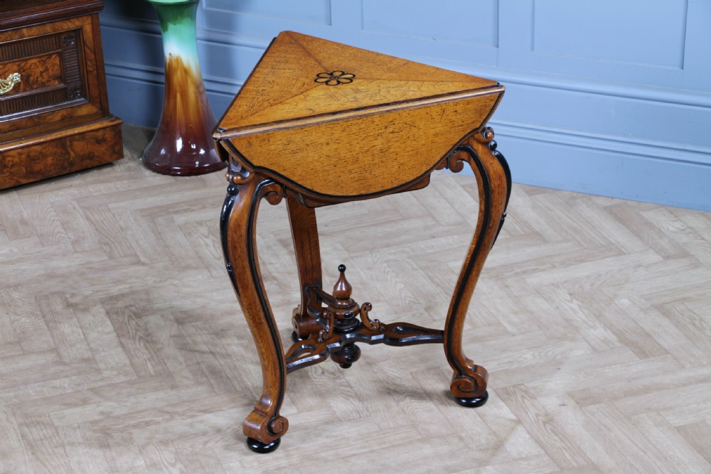 A Victorian Aesthetic Movement oak handkerchief table featuring ebonised accents, curved legs, and a triangular drop-leaf top with a central inlaid floral motif.