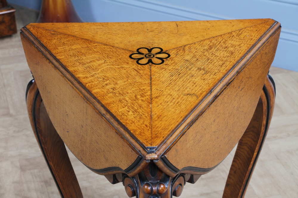 A Victorian Aesthetic Movement oak handkerchief table featuring ebonised accents, curved legs, and a triangular drop-leaf top with a central inlaid floral motif. View V