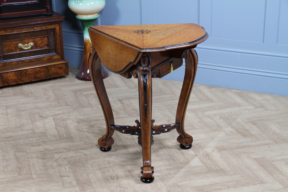 A Victorian Aesthetic Movement oak handkerchief table featuring ebonised accents, curved legs, and a triangular drop-leaf top with a central inlaid floral motif. View XIX