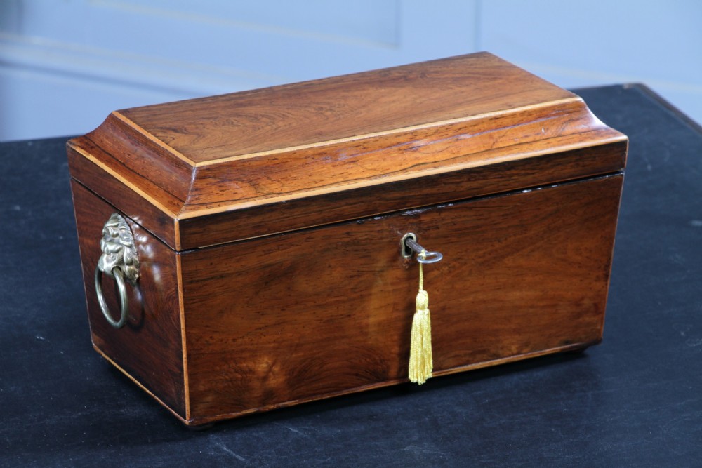 A Georgian rosewood sarcophagus-form tea caddy featuring stringing inlay, brass lion-mask side handles, and a keyhole with a tasselled key.