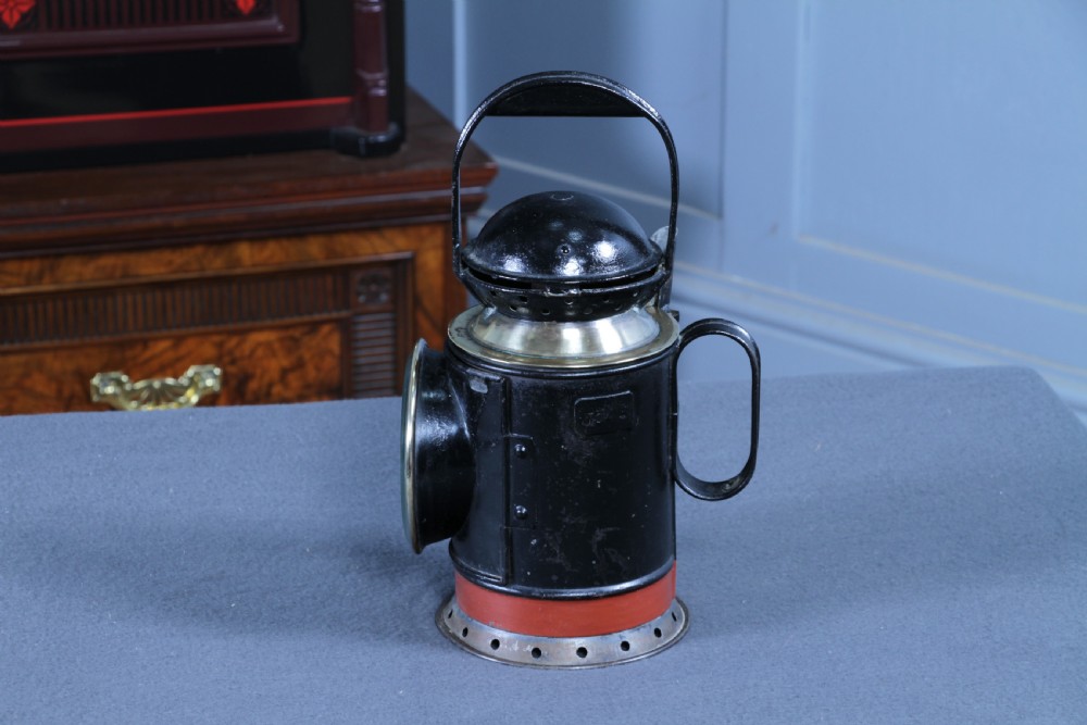 An antique black painted iron GWR railway hand lantern featuring a circular glass lens, top handle, brass accents, and a red painted band near the base. View V