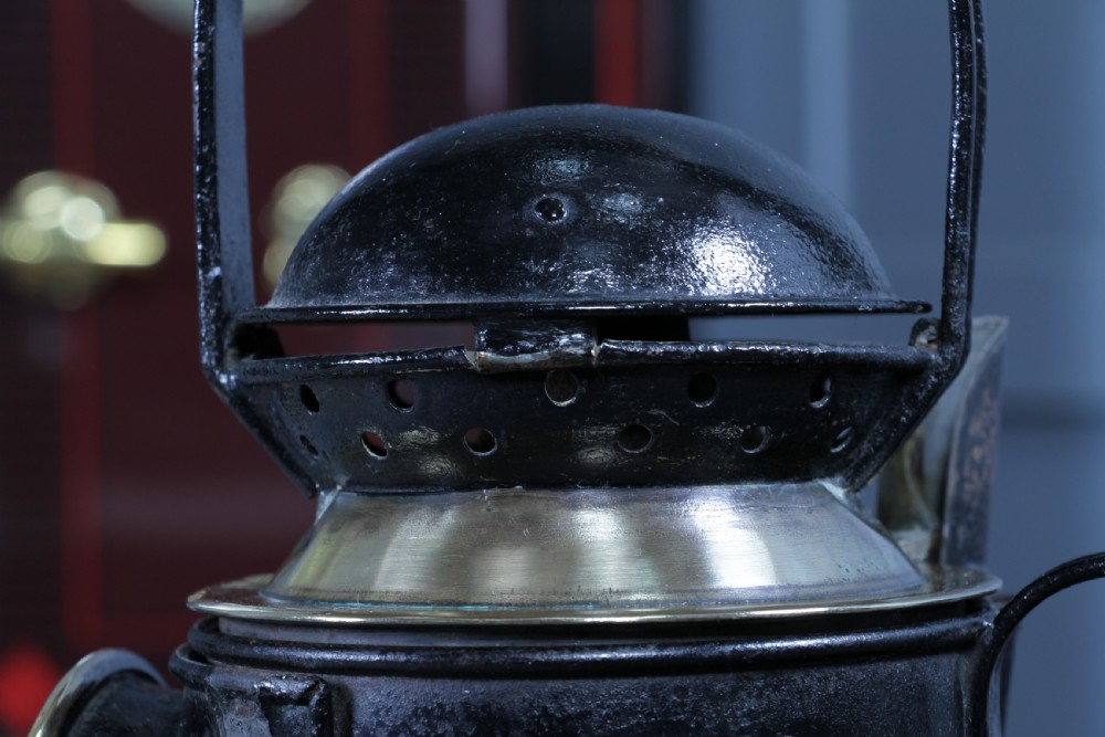 An antique black painted iron GWR railway hand lantern featuring a circular glass lens, top handle, brass accents, and a red painted band near the base. View VIII