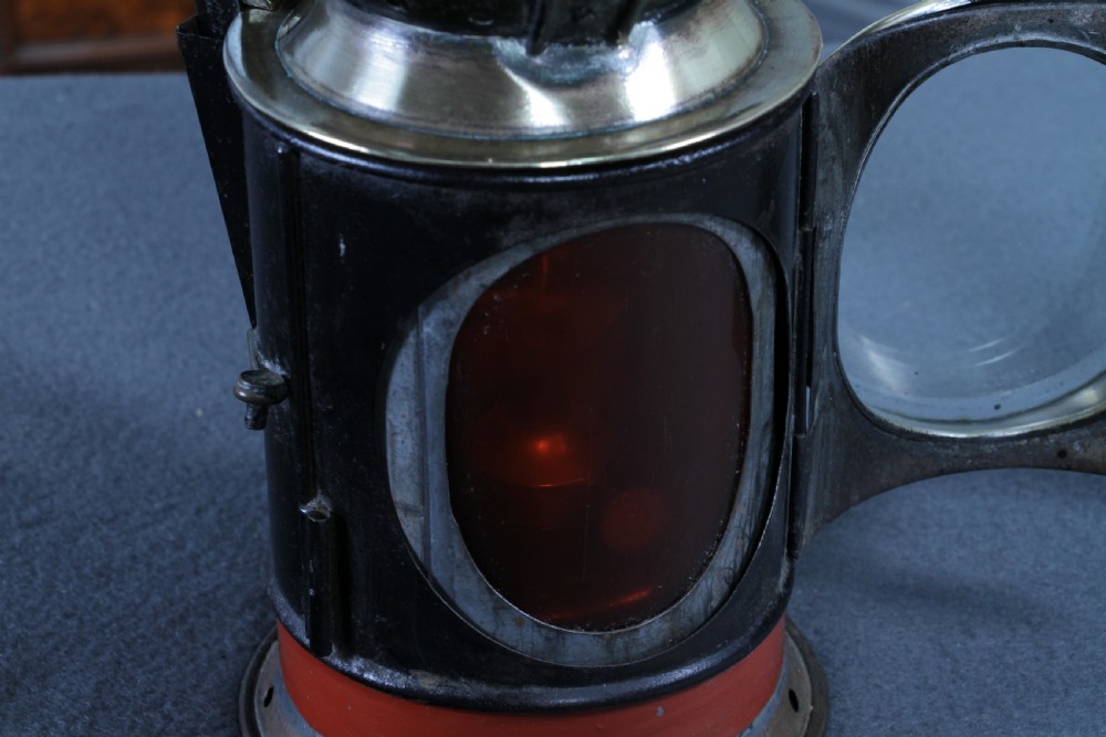 An antique black painted iron GWR railway hand lantern featuring a circular glass lens, top handle, brass accents, and a red painted band near the base. View X