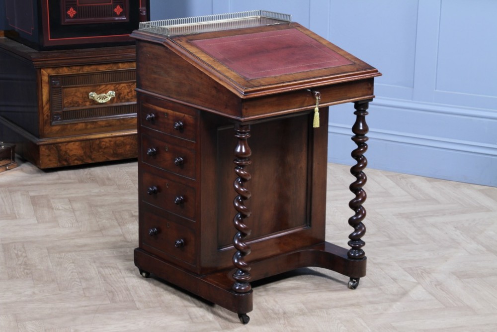 An early Victorian mahogany Davenport desk featuring a sloped writing surface with red leather inset, brass top gallery, barley twist front supports, and four side drawers.