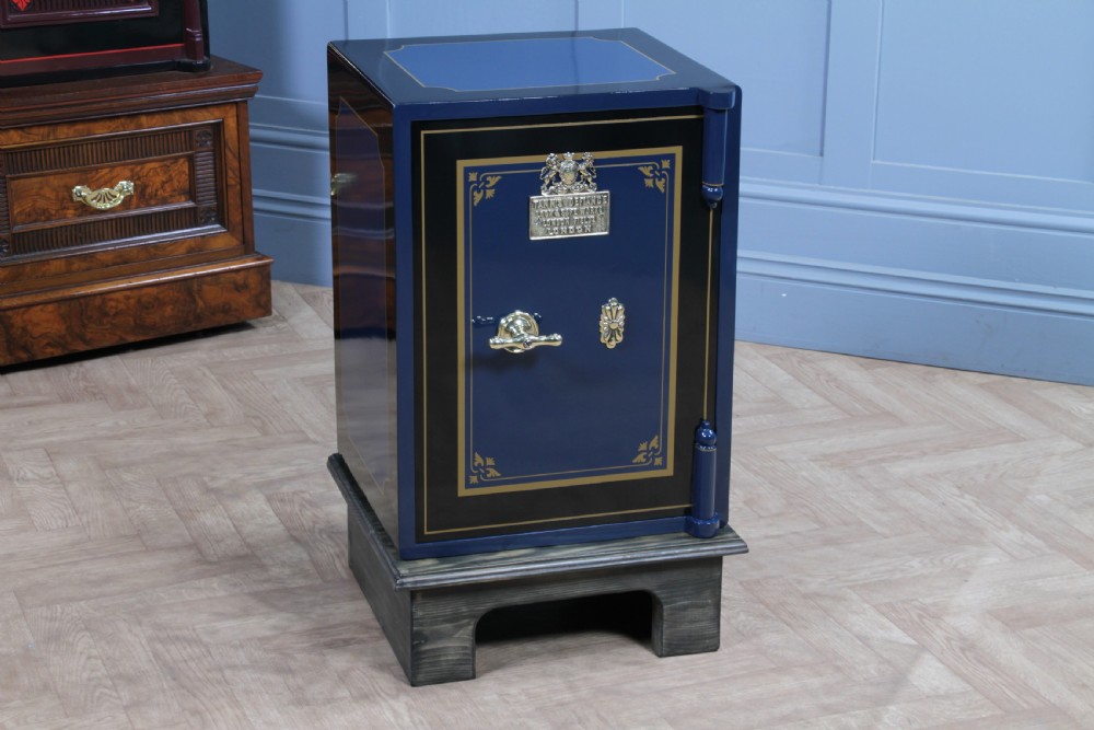 A restored Victorian navy blue and black cast iron safe featuring polished brass hardware and a makers plaque, raised on a dark wooden plinth.