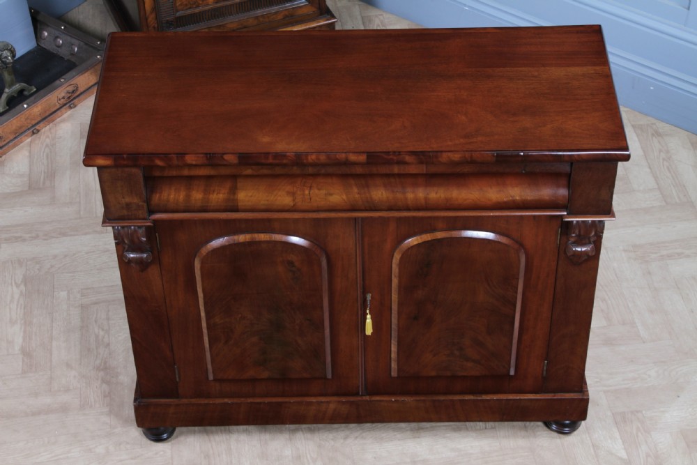 A Victorian flame mahogany two-door sideboard featuring a cushion drawer, arched panel doors, carved corbels, and turned bun feet. View V