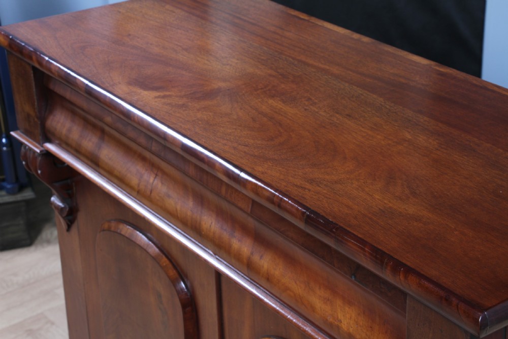 A Victorian flame mahogany two-door sideboard featuring a cushion drawer, arched panel doors, carved corbels, and turned bun feet. View XV