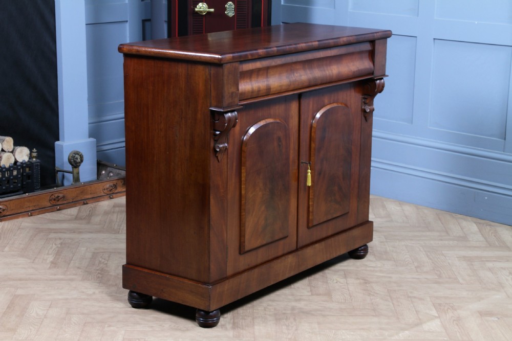 A Victorian flame mahogany two-door sideboard featuring a cushion drawer, arched panel doors, carved corbels, and turned bun feet. View XIX