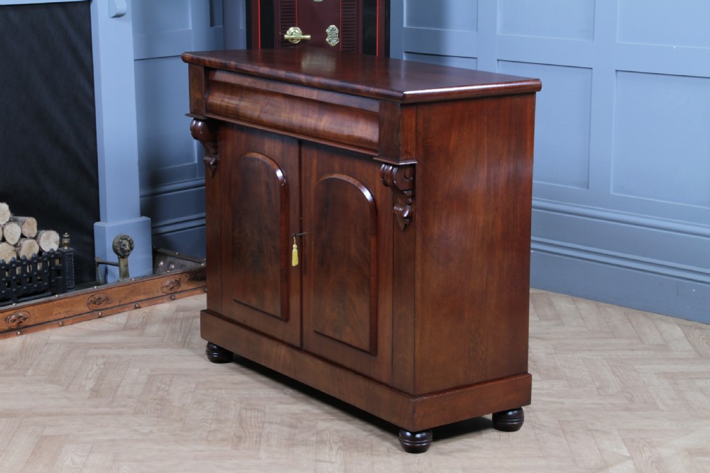 A Victorian flame mahogany two-door sideboard featuring a cushion drawer, arched panel doors, carved corbels, and turned bun feet. View XX