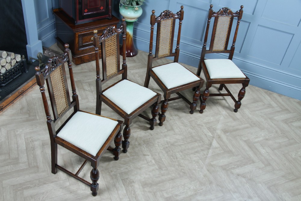 A set of four dark oak Jacobean Revival dining chairs featuring woven cane backs, carved crests, turned legs, and cream fabric seats. View II