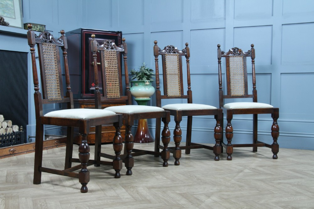 A set of four dark oak Jacobean Revival dining chairs featuring woven cane backs, carved crests, turned legs, and cream fabric seats. View IV