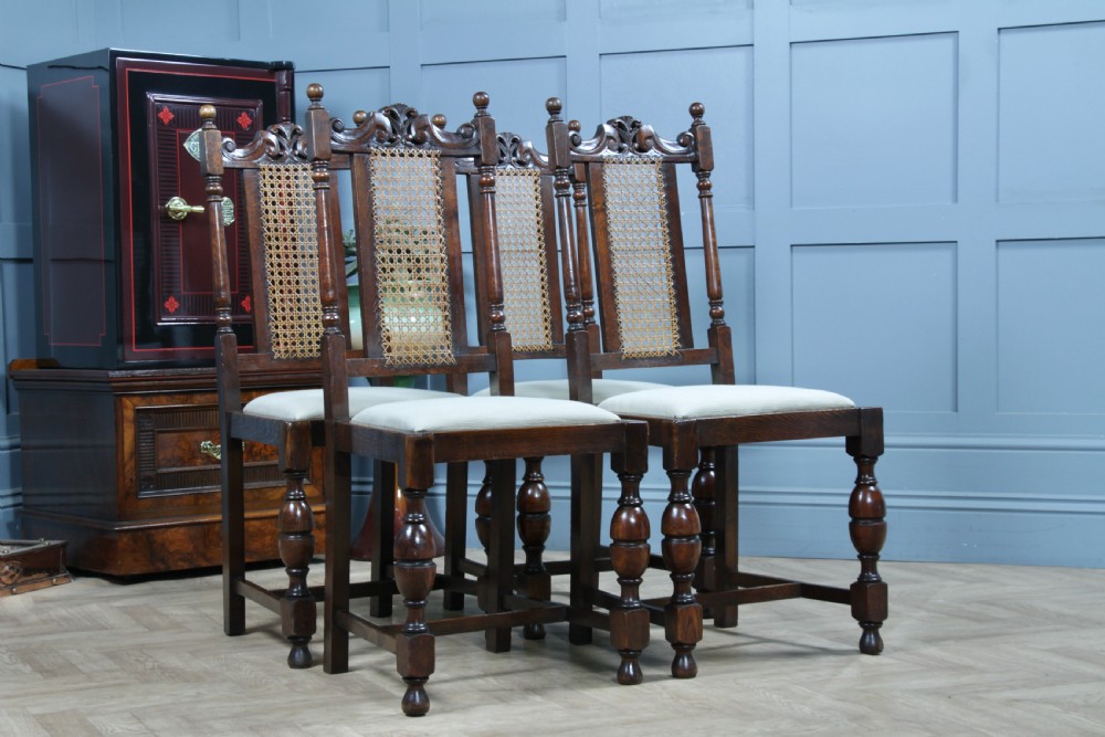 A set of four dark oak Jacobean Revival dining chairs featuring woven cane backs, carved crests, turned legs, and cream fabric seats. View V