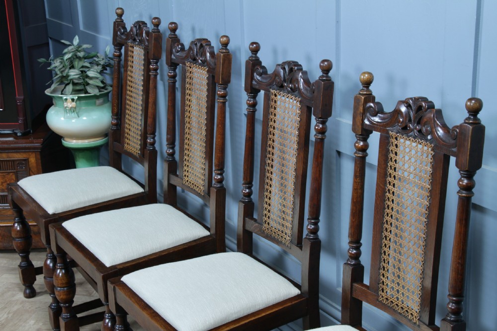 A set of four dark oak Jacobean Revival dining chairs featuring woven cane backs, carved crests, turned legs, and cream fabric seats. View VI