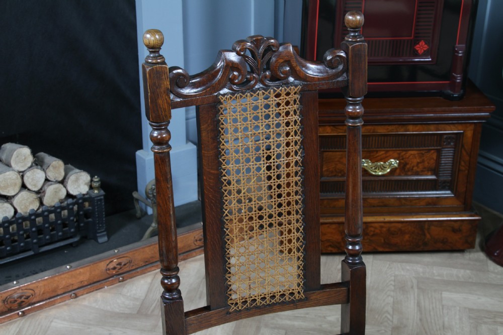 A set of four dark oak Jacobean Revival dining chairs featuring woven cane backs, carved crests, turned legs, and cream fabric seats. View VII