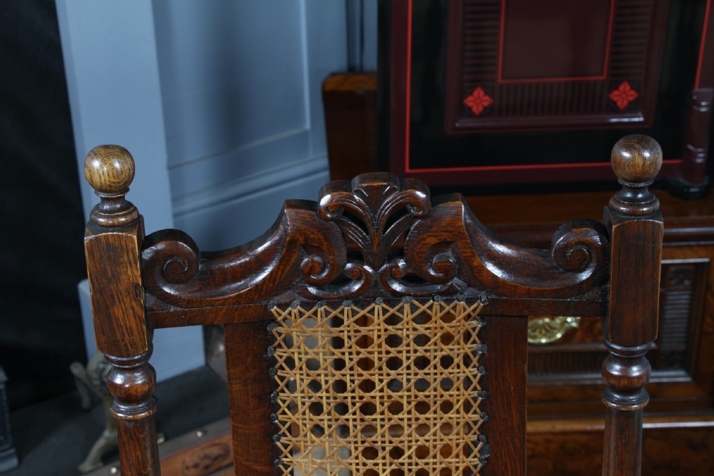 A set of four dark oak Jacobean Revival dining chairs featuring woven cane backs, carved crests, turned legs, and cream fabric seats. View VIII