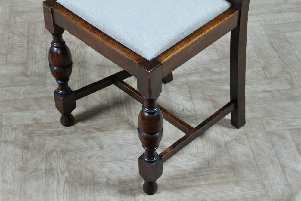 A set of four dark oak Jacobean Revival dining chairs featuring woven cane backs, carved crests, turned legs, and cream fabric seats. View IX