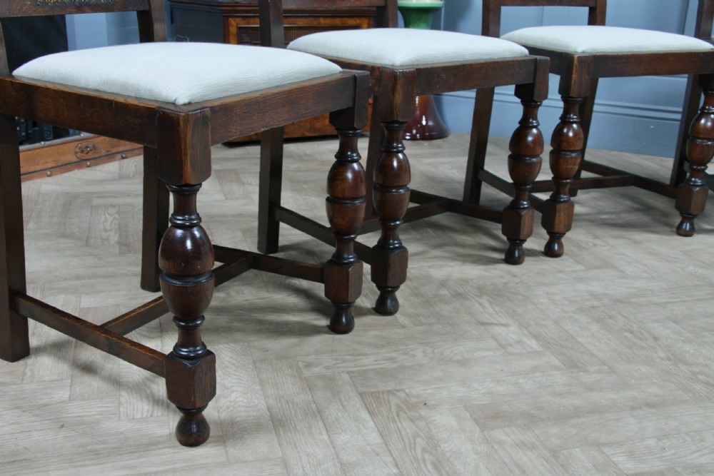 A set of four dark oak Jacobean Revival dining chairs featuring woven cane backs, carved crests, turned legs, and cream fabric seats. View X