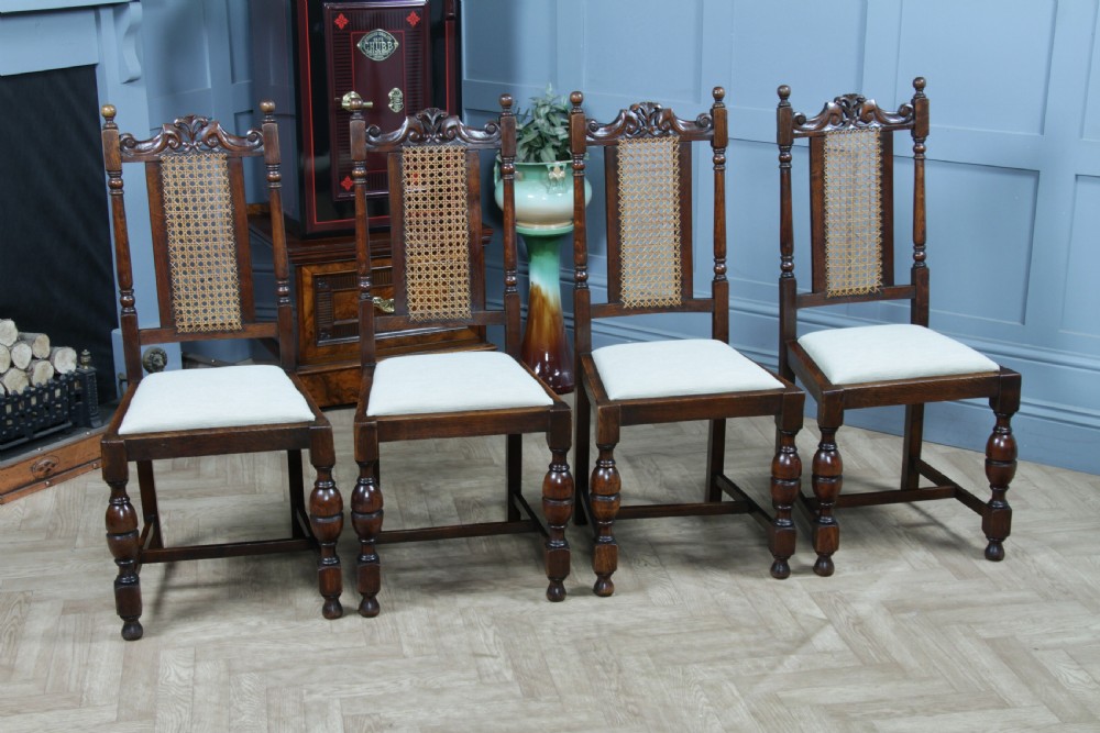 A set of four dark oak Jacobean Revival dining chairs featuring woven cane backs, carved crests, turned legs, and cream fabric seats. View XIII