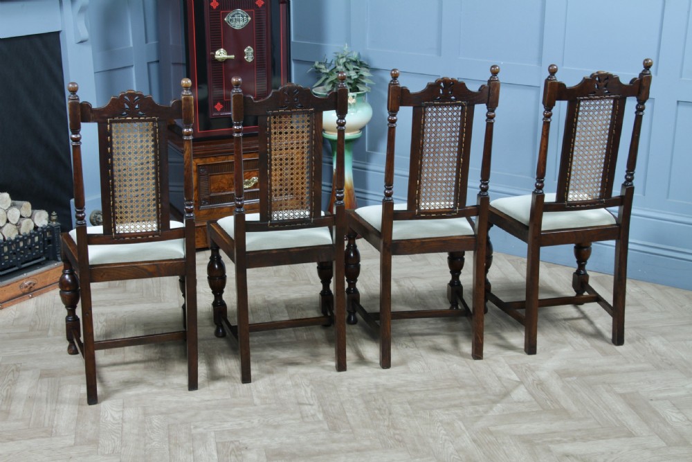 A set of four dark oak Jacobean Revival dining chairs featuring woven cane backs, carved crests, turned legs, and cream fabric seats. View XV