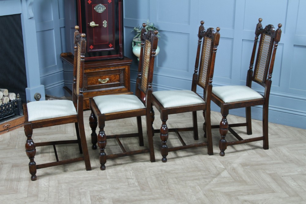 A set of four dark oak Jacobean Revival dining chairs featuring woven cane backs, carved crests, turned legs, and cream fabric seats. View XVI