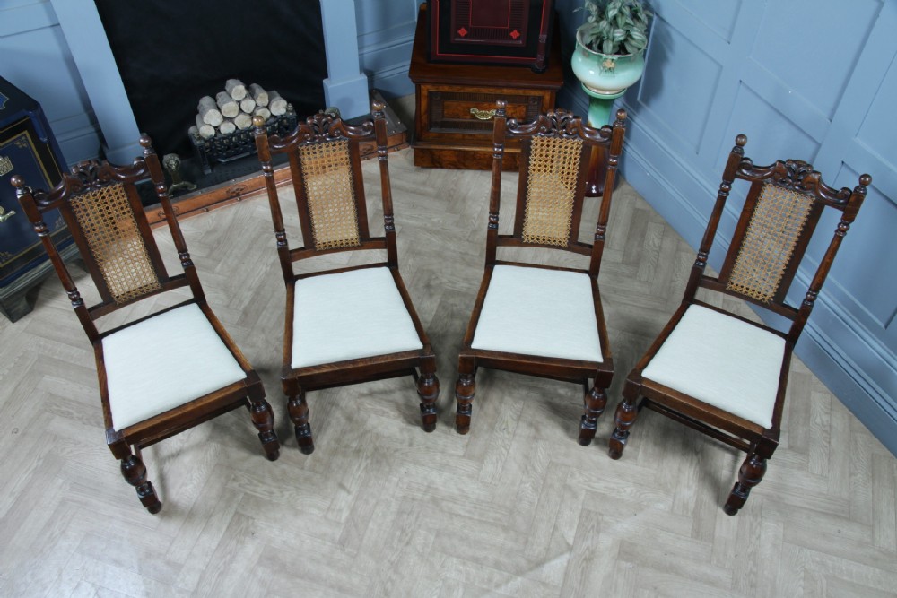 A set of four dark oak Jacobean Revival dining chairs featuring woven cane backs, carved crests, turned legs, and cream fabric seats. View XVII
