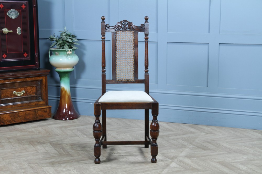 A set of four dark oak Jacobean Revival dining chairs featuring woven cane backs, carved crests, turned legs, and cream fabric seats. View XVIII
